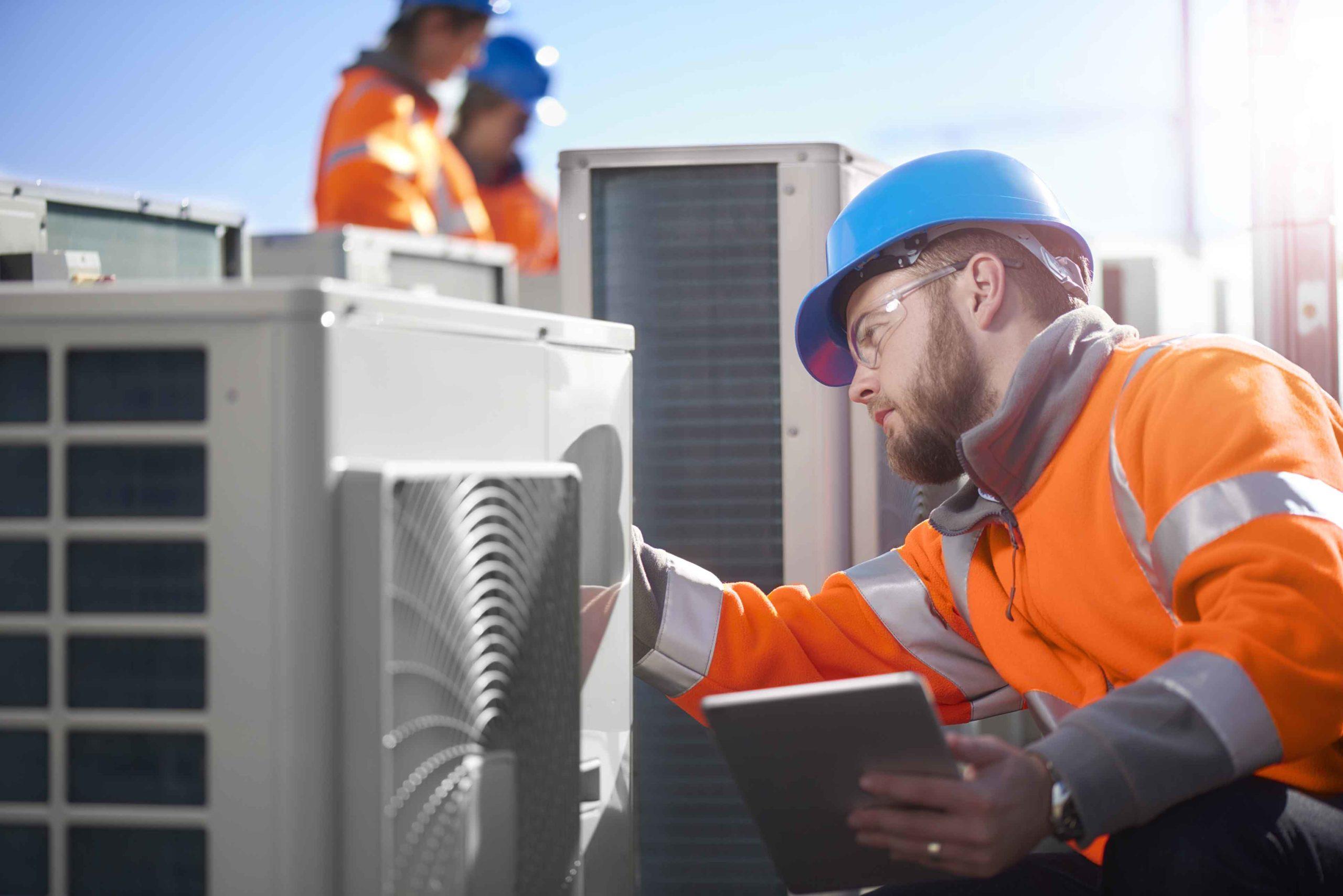 Engineer in orange overalls working on air conditioning unit while holding iPad tablet computer