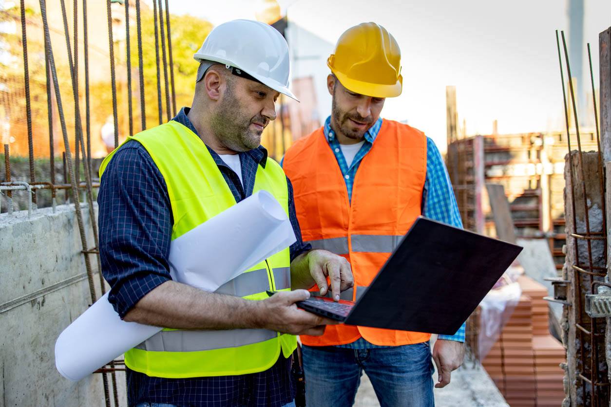 Two construction workers wearing high visibility jackets and ppe with one holding laptop and pointing to screen in working area